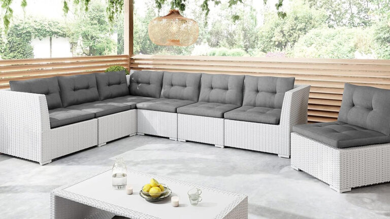 Nowoczesne patio na świeżym powietrzu z białą wiklinową sofą segmentową z szarymi poduszkami, pasującym stolikiem kawowym z owocami, szklankami i świecami, pod plecioną oprawą oświetleniową i otoczone zielenią.