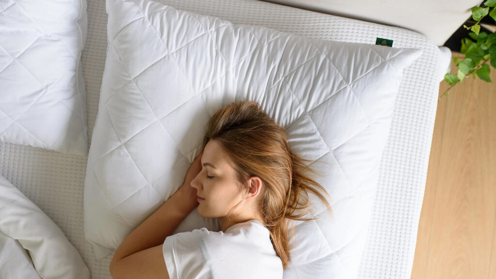 Kobieta o jasnobrązowych włosach śpi na boku, opierając głowę na dużej białej poduszce. Leży na białym łóżku, częściowo przykryta białym kocem. W rogu widoczna jest zielona roślina.